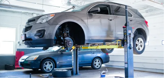 SUV raised on a hydraulic lift in an auto repair shop with another car parked nearby.