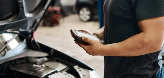 Mechanic holding a device while standing next to a car with an open hood.