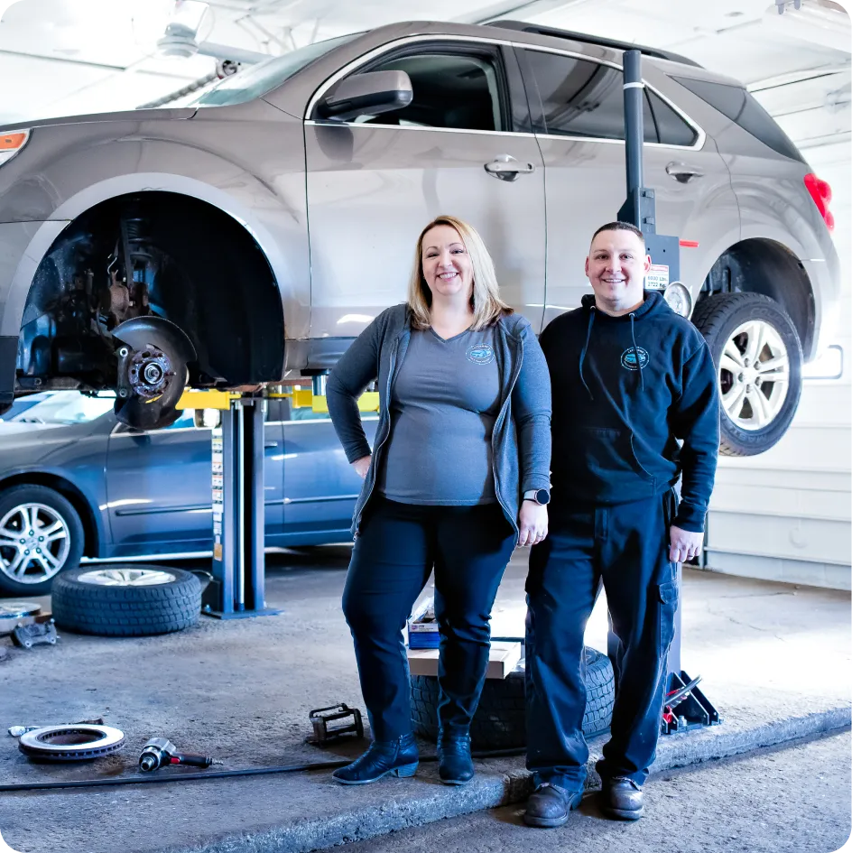 careers, auto & truck services in Westfield, MA at LaFlamme's Auto & Truck Services. A silver SUV is raised on a hydraulic lift with Sarah and Wayne LaFlamme standing in front of it inside a repair shop.
